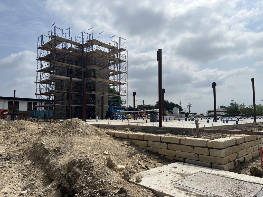 Image shows construction site for the new Cairn Point Cameron. A tall concrete elevator shaft surrounded by metal scaffolding can be seen in the background. In the foreground, there is dirt; also plumbing pipes and metal columns are shown coming out of the concrete foundation.
