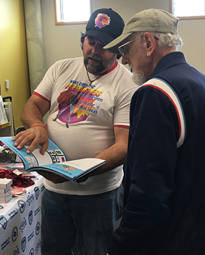 younger man wearing a hat shows an elderly man a manual on emergency preparedness.