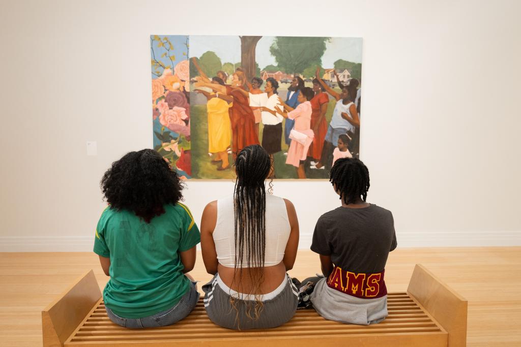 Three people sitting on a bench looking at a painting by Riley Holloway at the George Washington Carver Museum in Austin, TX.