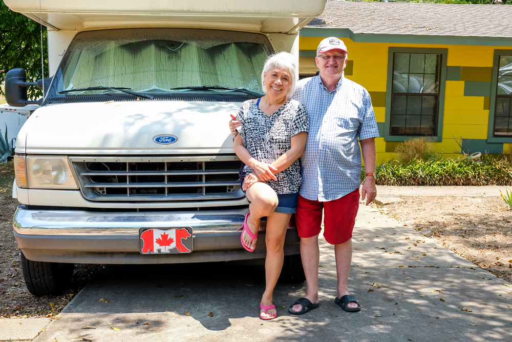 A couple standing in front of an RV smiling. The RV is parked in the driveway of a yellow one-story hourse. The license plate is canadian. 