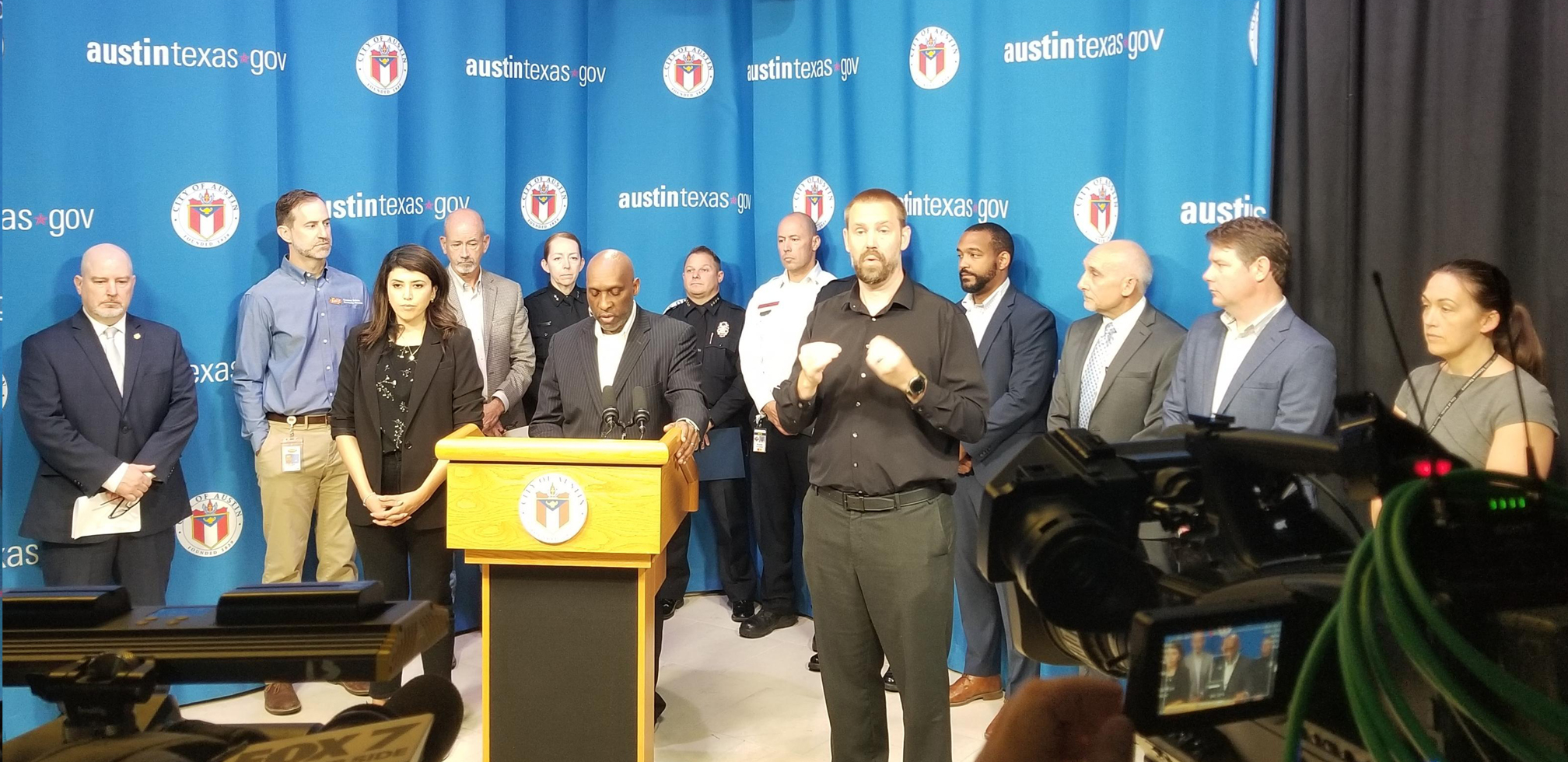 image of black man speaking behind podium at a press conference. A group of city officials stand behind him and an ASL interpreter signs next to him.