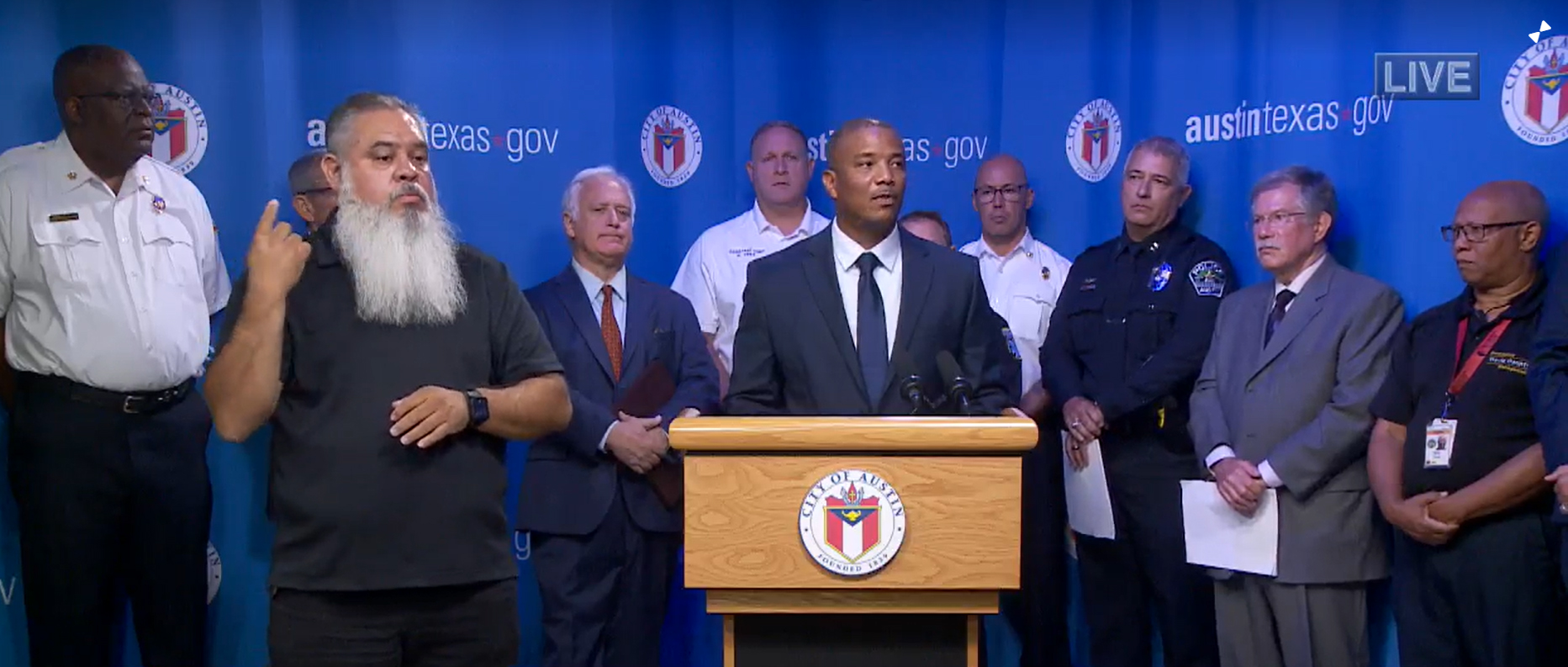 Multiple city officials standing behind a podium in front of a City of Austin backdrop. A black male stands at podium speaking.