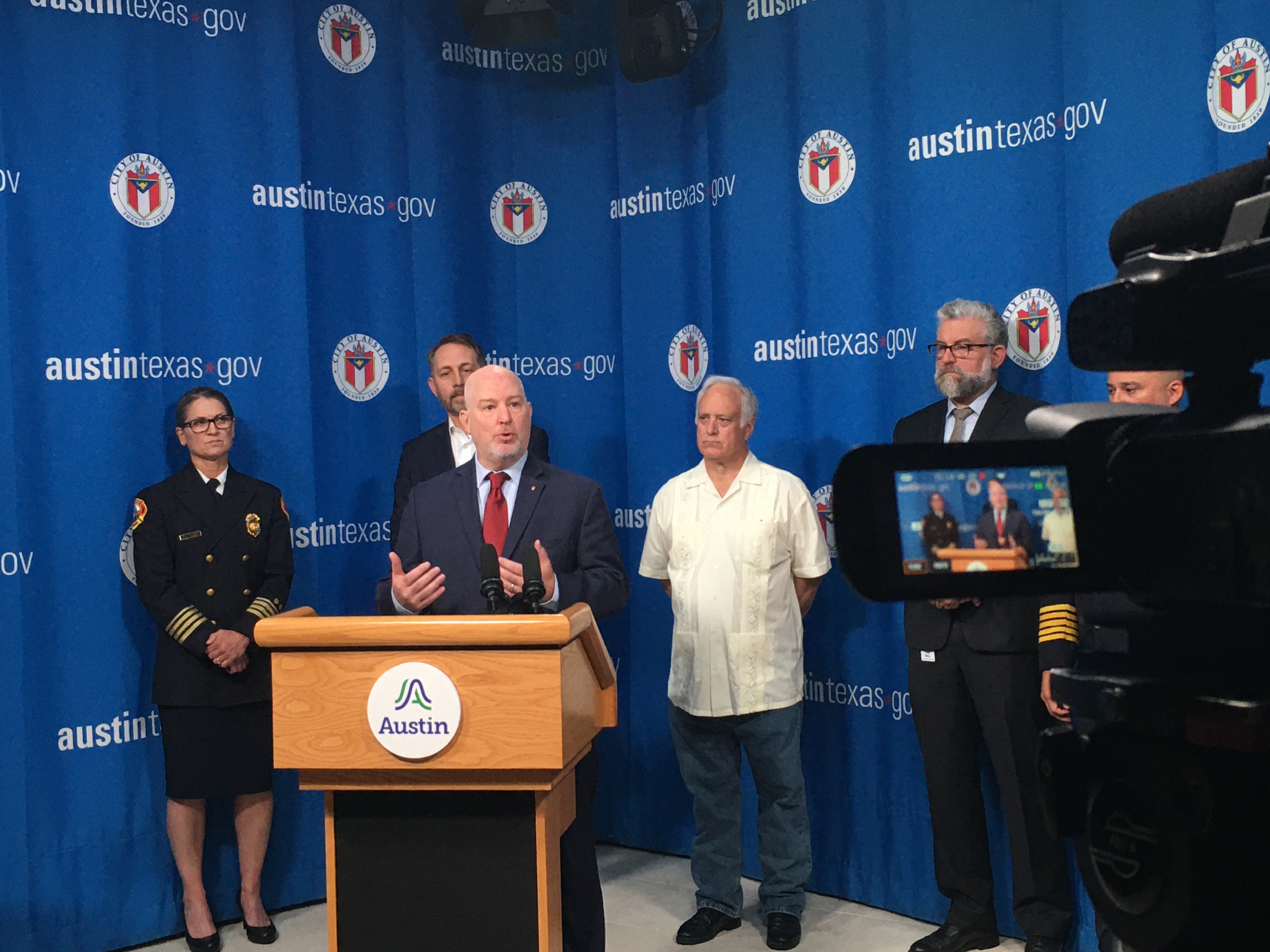 Middle-aged man in blue suit stands behind podium speaking at a press conference. Other government officials stand behind him in front of a curtain with the City of Austin seal and logo on it.