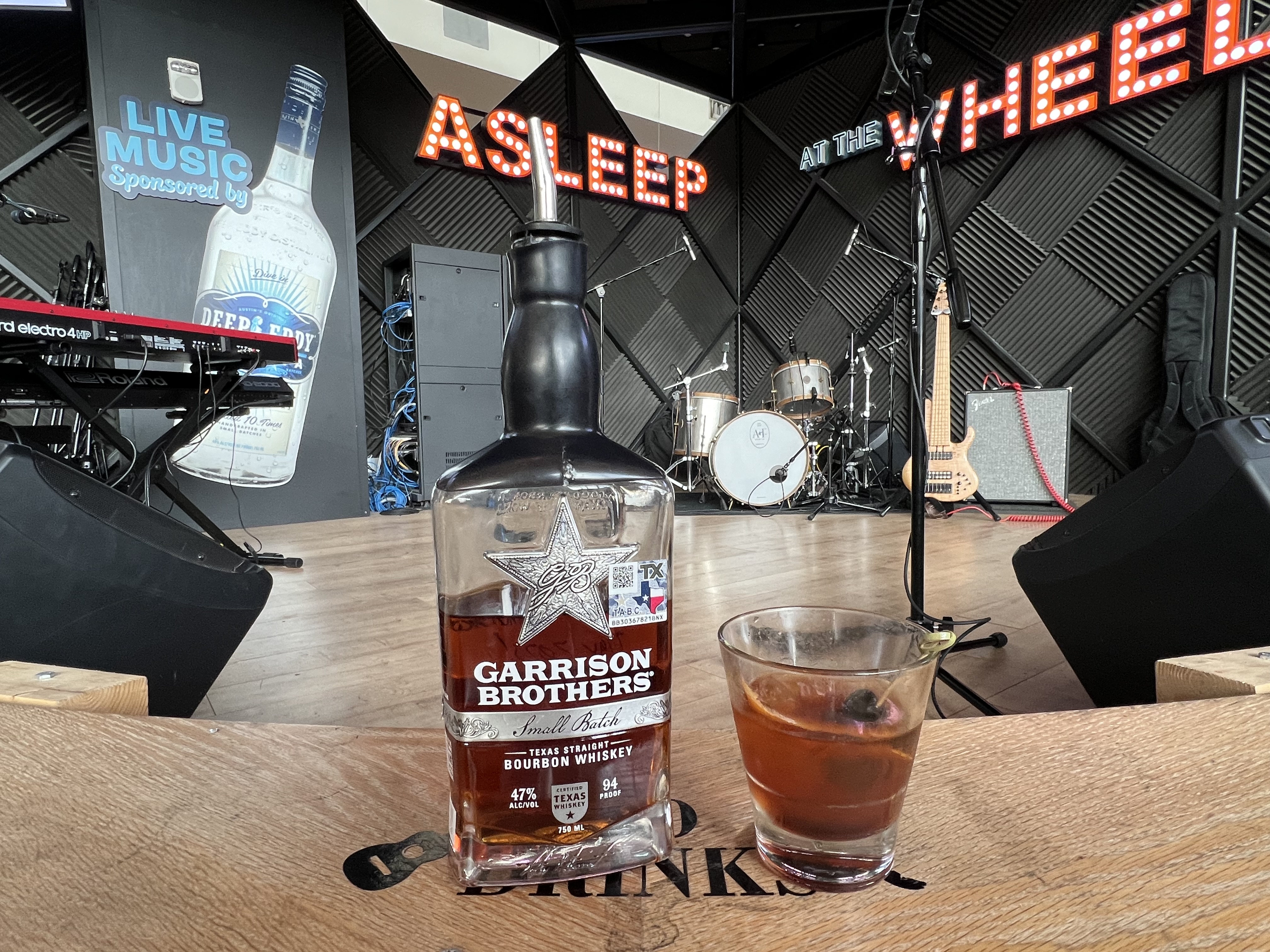 A close-up photo of the Garrison Bros whiskey and an Old Fashioned cocktail. The Asleep at the Wheel stage at the Saxon Pub is in the background.