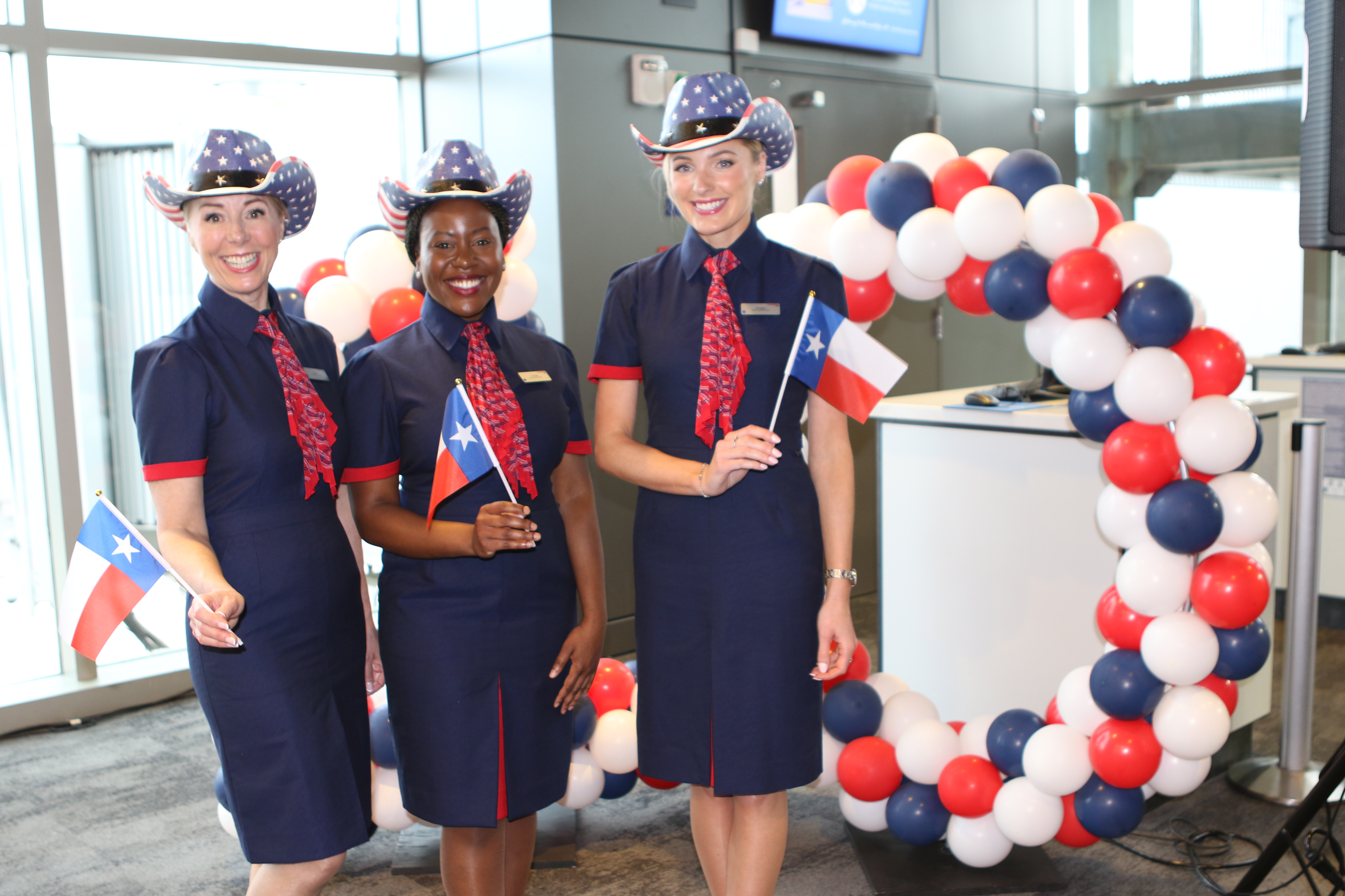 Photo of 3 British Airways flight attendants standing and posing for a photo at the AUS/British Airways 10 year anniversary celebration. They're holding tiny Texas flags.
