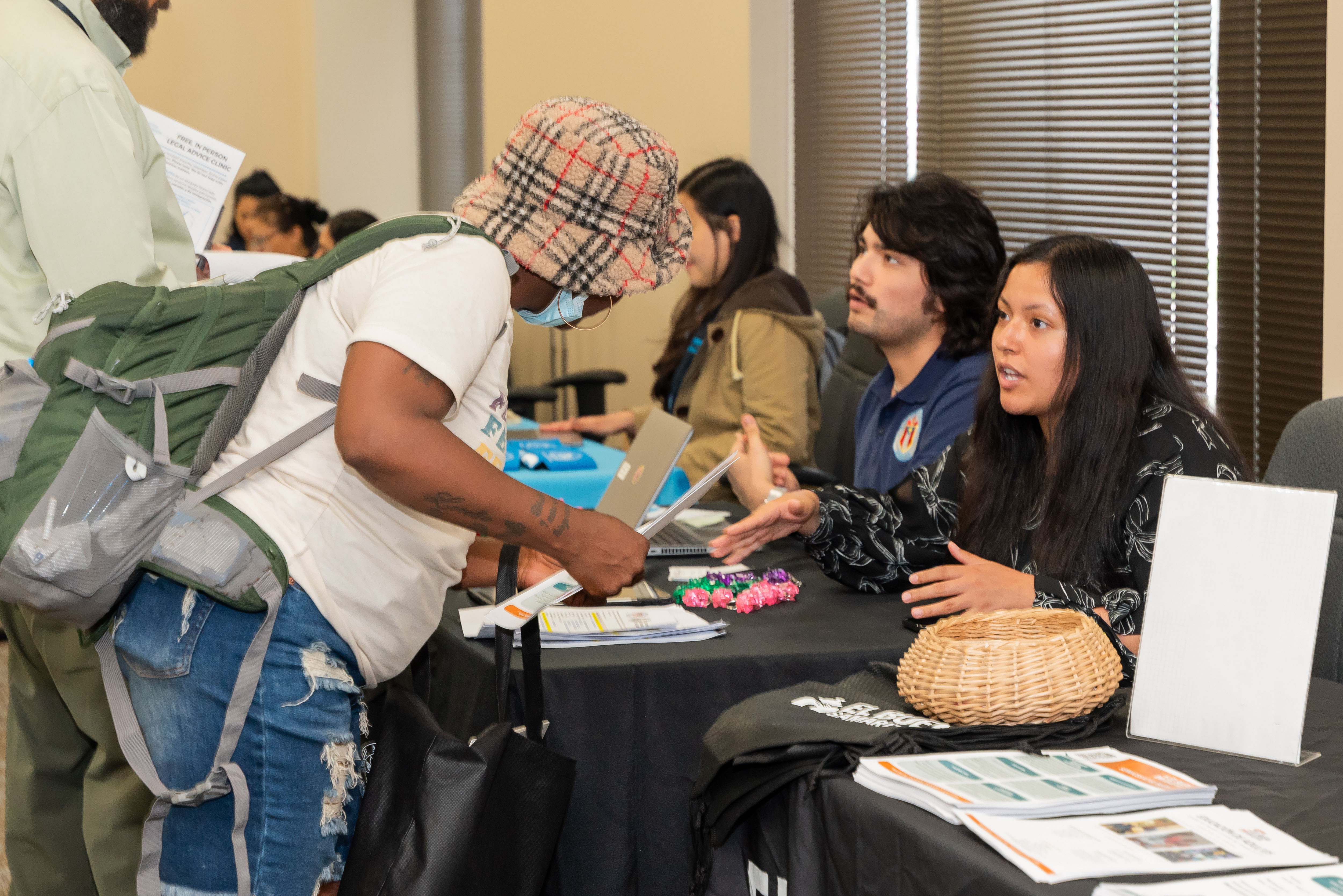 Two city of Austin employees in the foreground and seated a table with a black tablecloth, greet Austin residents and provide them with pamphlets and flyers on services available to the community.