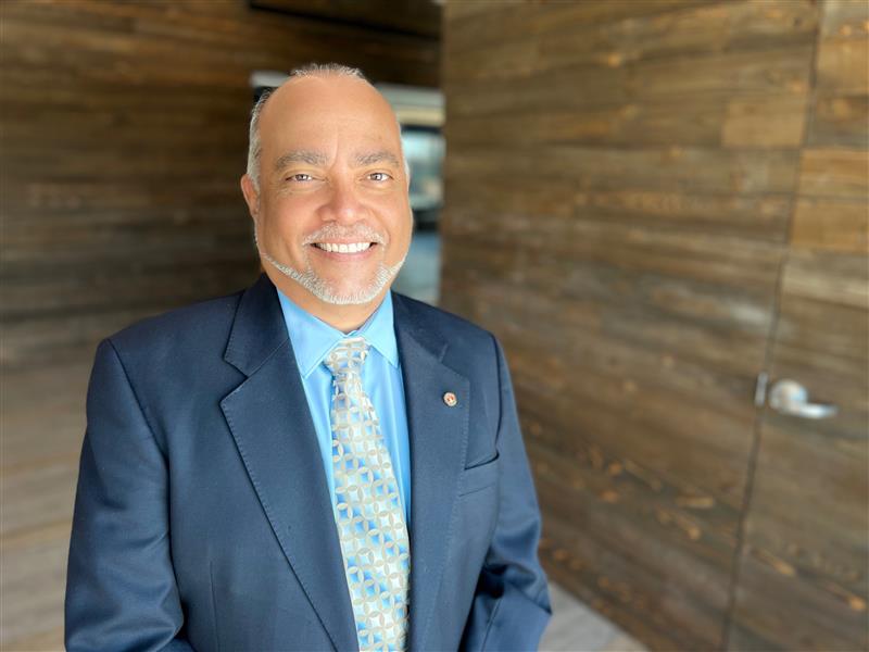 Austin Code Director Jose Roig stands in an hallway in the Austin Code offices. Mr. Roig is wearing a blue suit with a striped necktie. He is smiling and looking directly into the camera.