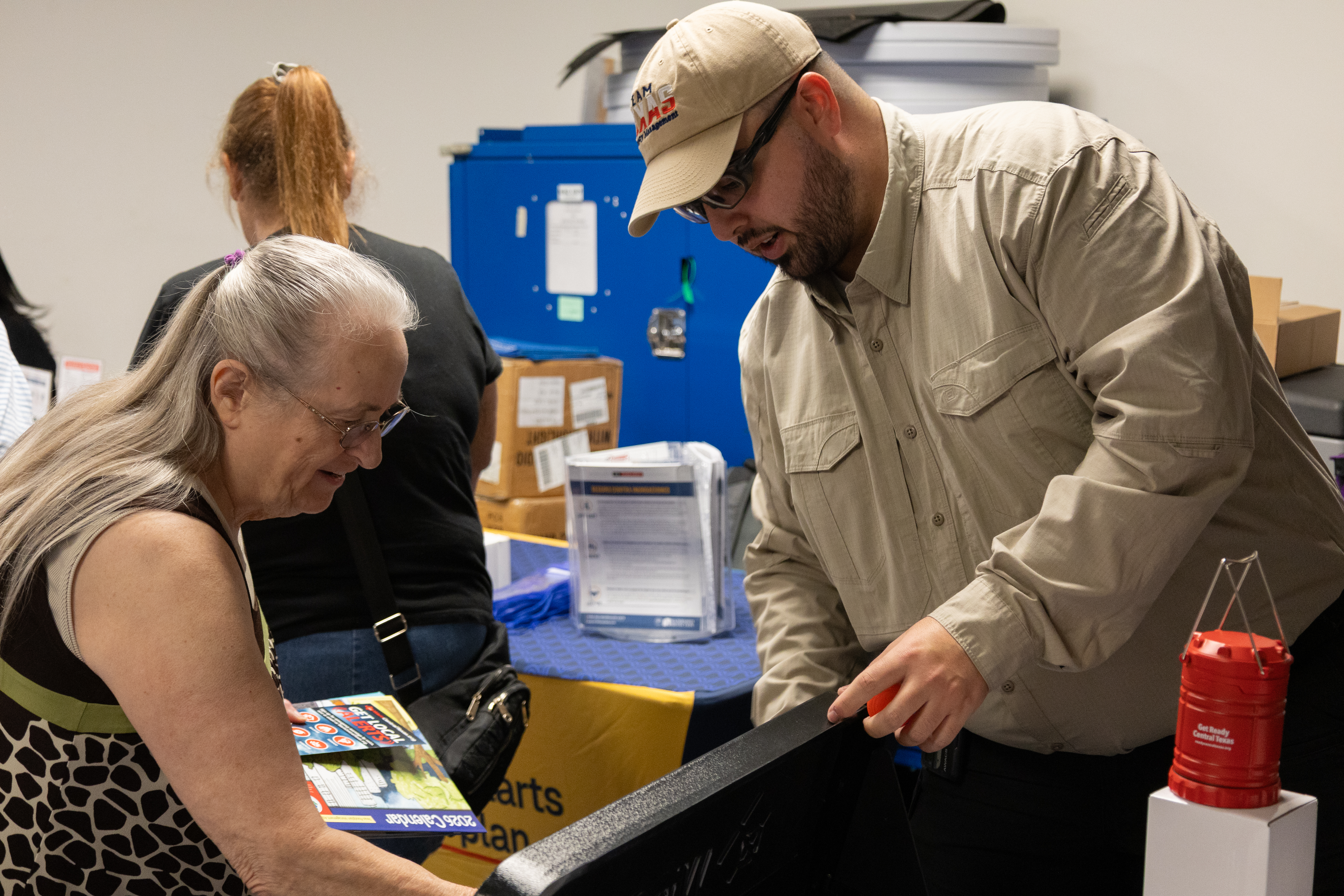 an elderly woman talks to a man wearing a ballcap and glasses at a public event for emergency preparedness.