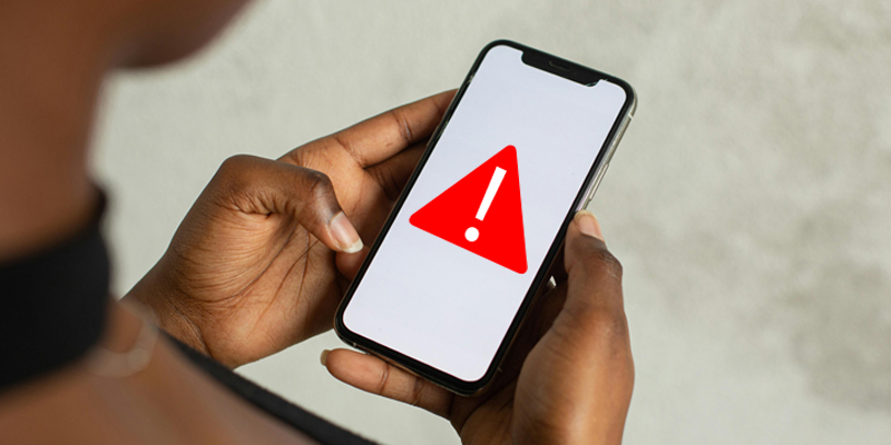 cell phone alert image of an African-American woman's hands holding a cell phone. The phone has an image of a red triangle and exclamation mark to indicate an alert.