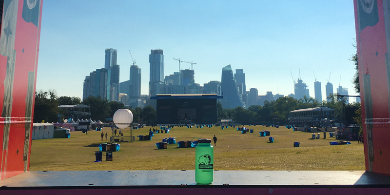 A photo showing an Austin HSEM water bottle in front of the ACL festival with the Austin skyline in the background.