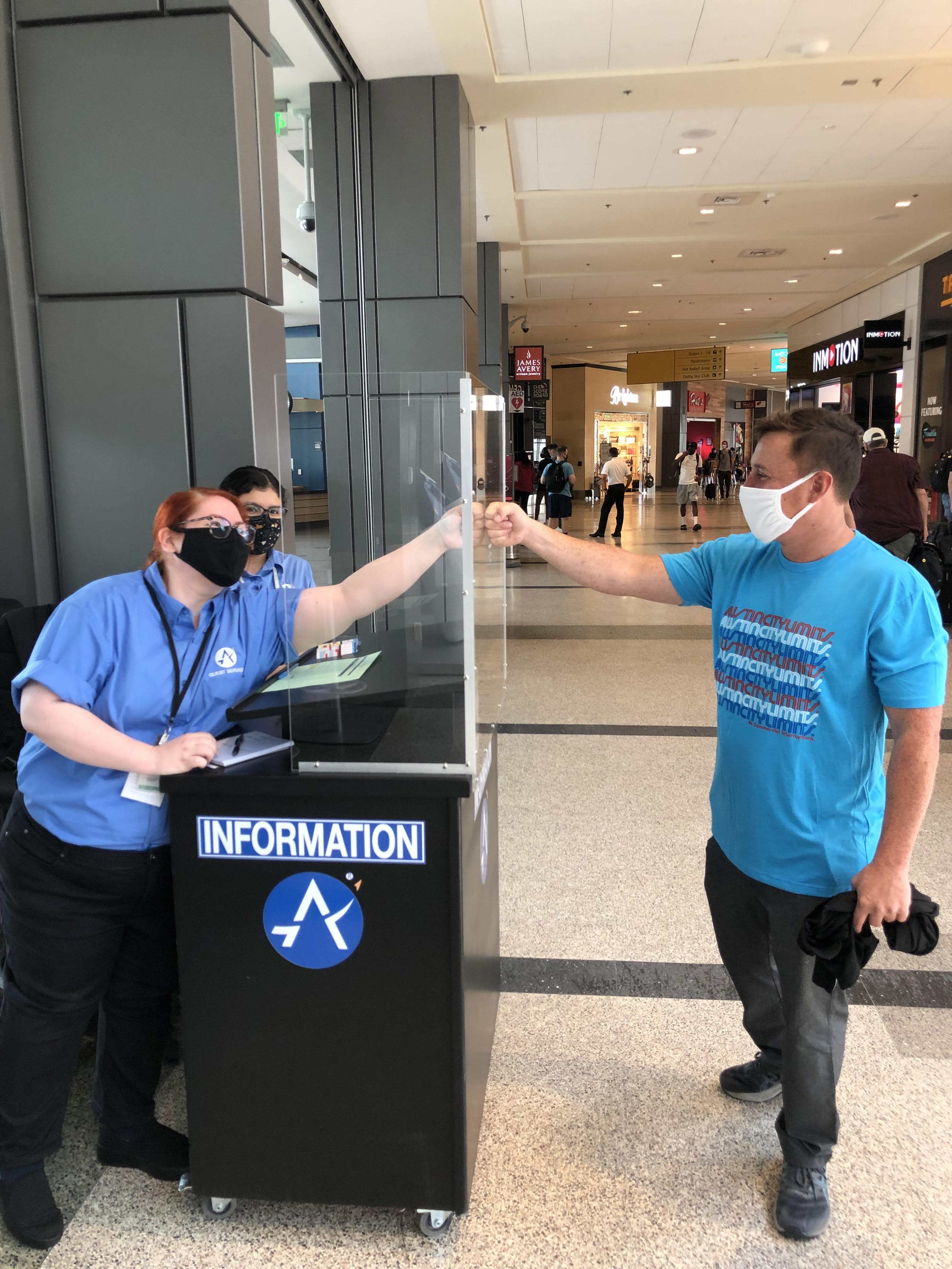 A passenger fist bumps an airport employee from opposite sides of a plexiglass safety barrier.