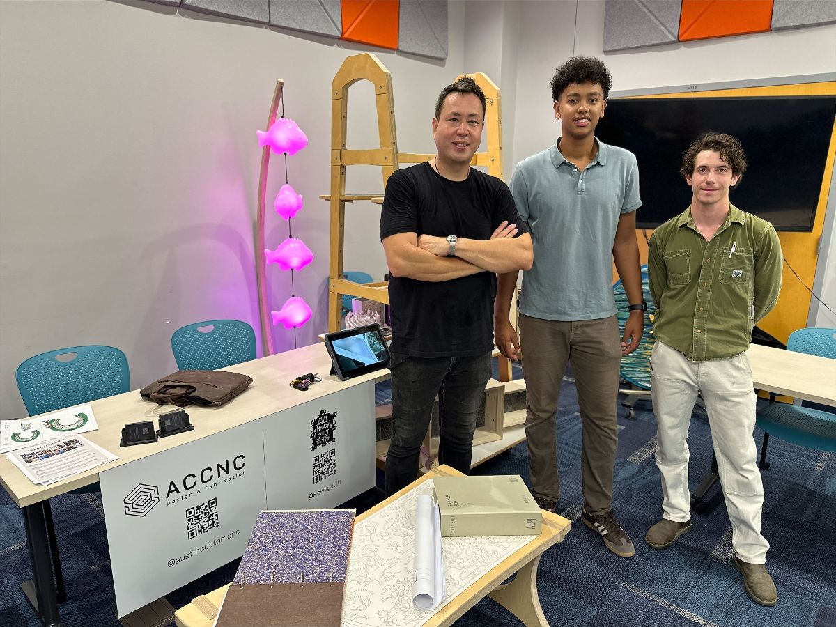 Austin Custom CNC and HowdyBuilt attend the inaugural Public Art Makers Expo Three men stand and smile for a photo behind a display table at an event. The table has design samples, booklets, and a tablet, with a banner showing the ACCNC Designs & Fabrication logo and QR codes. Behind them is a wooden display structure and a lamp with pink glowing lights shaped like clouds.