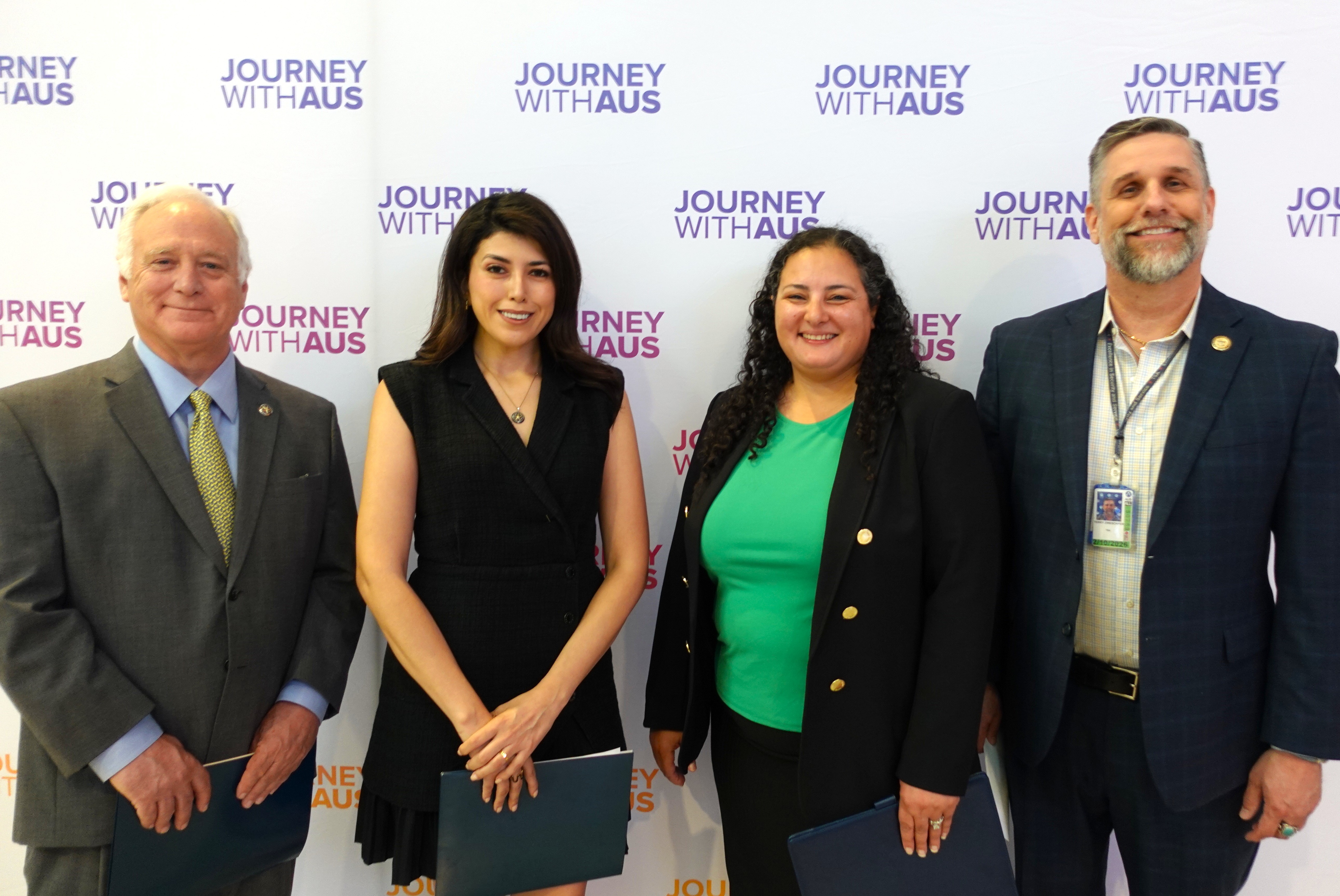 From left to right: Austin Mayor Kirk Watson, Austin Mayor Pro Tem Vanessa Fuentes, AUS CEO Ghizlane Badawi, and TSA Deputy Assistant Federal Security Director Terry Zweschper pose for a photo in front of a "Journey With AUS" branded backdrop at the Atrium Infill event. They are holding blue folders and are dressed in professional attire, smiling confidently for the camera.
