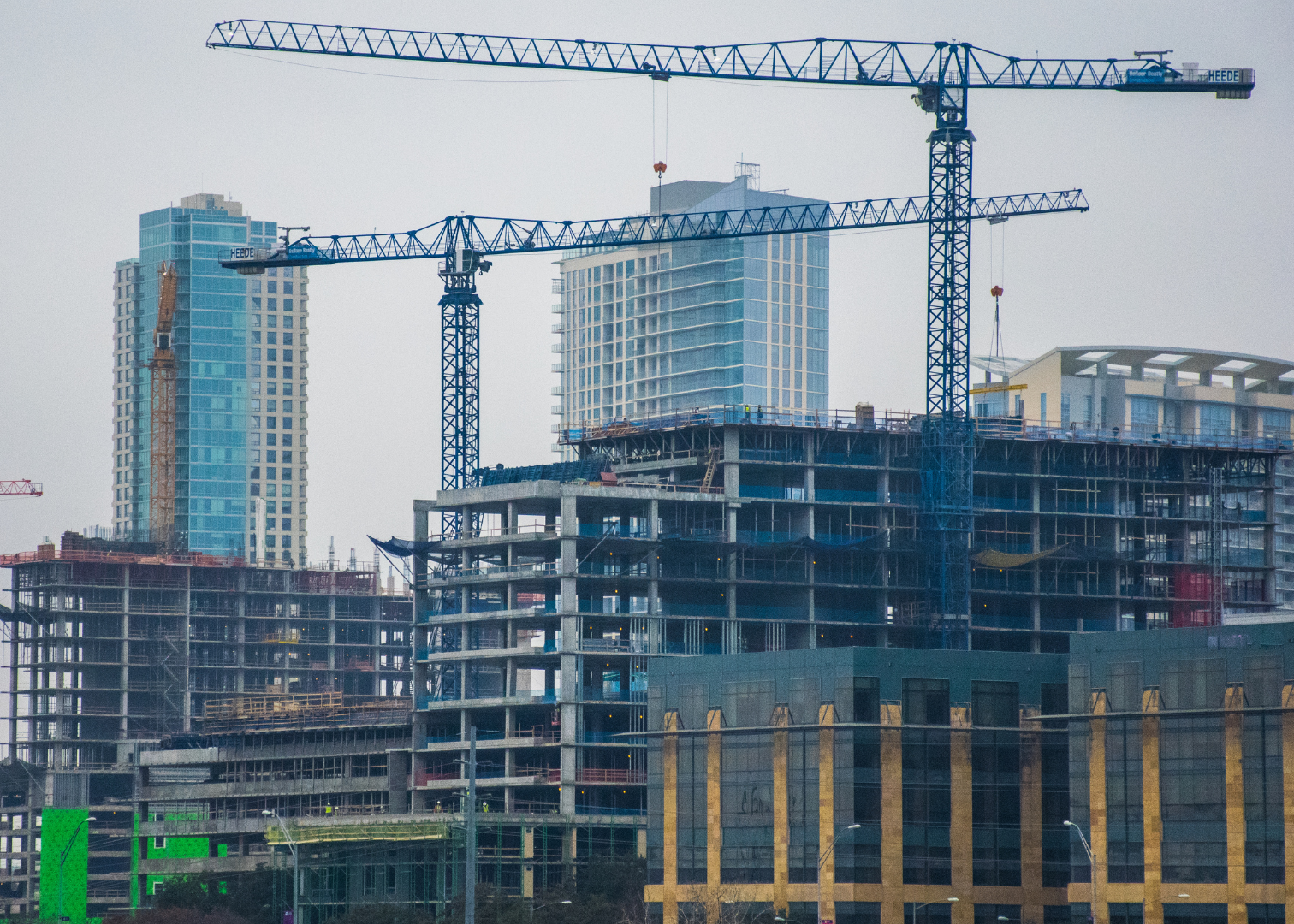 Portion of the Austin skyline with construction machines building a new skyscraper. Center of the frame is a crane over a building with two other buildings in the background. The sky is light gray.