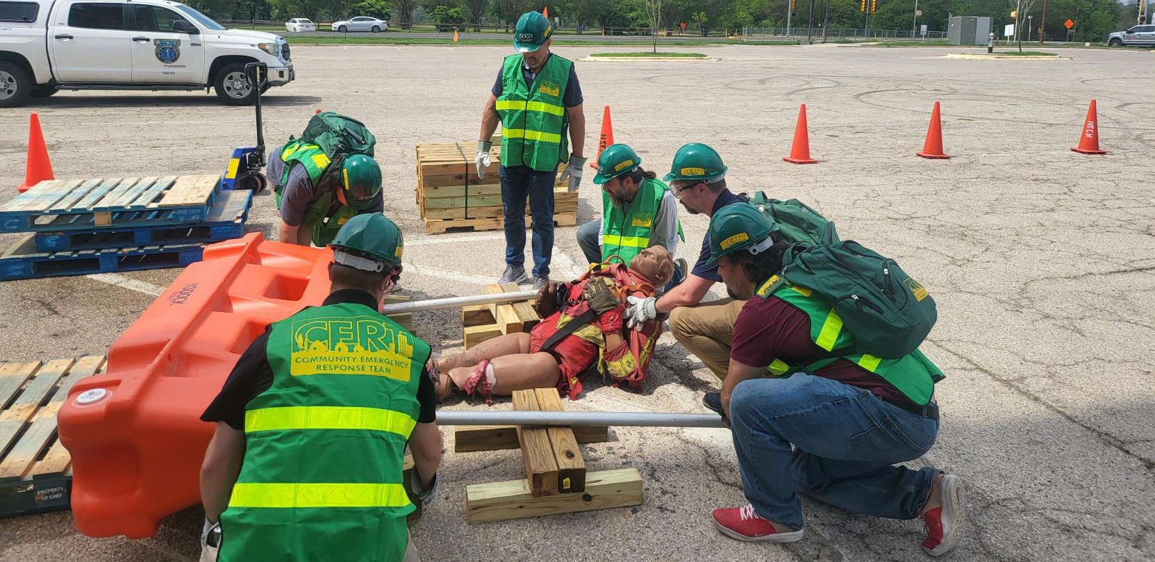 CERT exercise photo1 A group of individuals wearing Community Emergency Response Team vests and personal protective gear including gloves, helmets, and goggles use wood and pipes as "cribbing" to create a lever and lift a heavy object off of a dummy in a simulated emergency response.