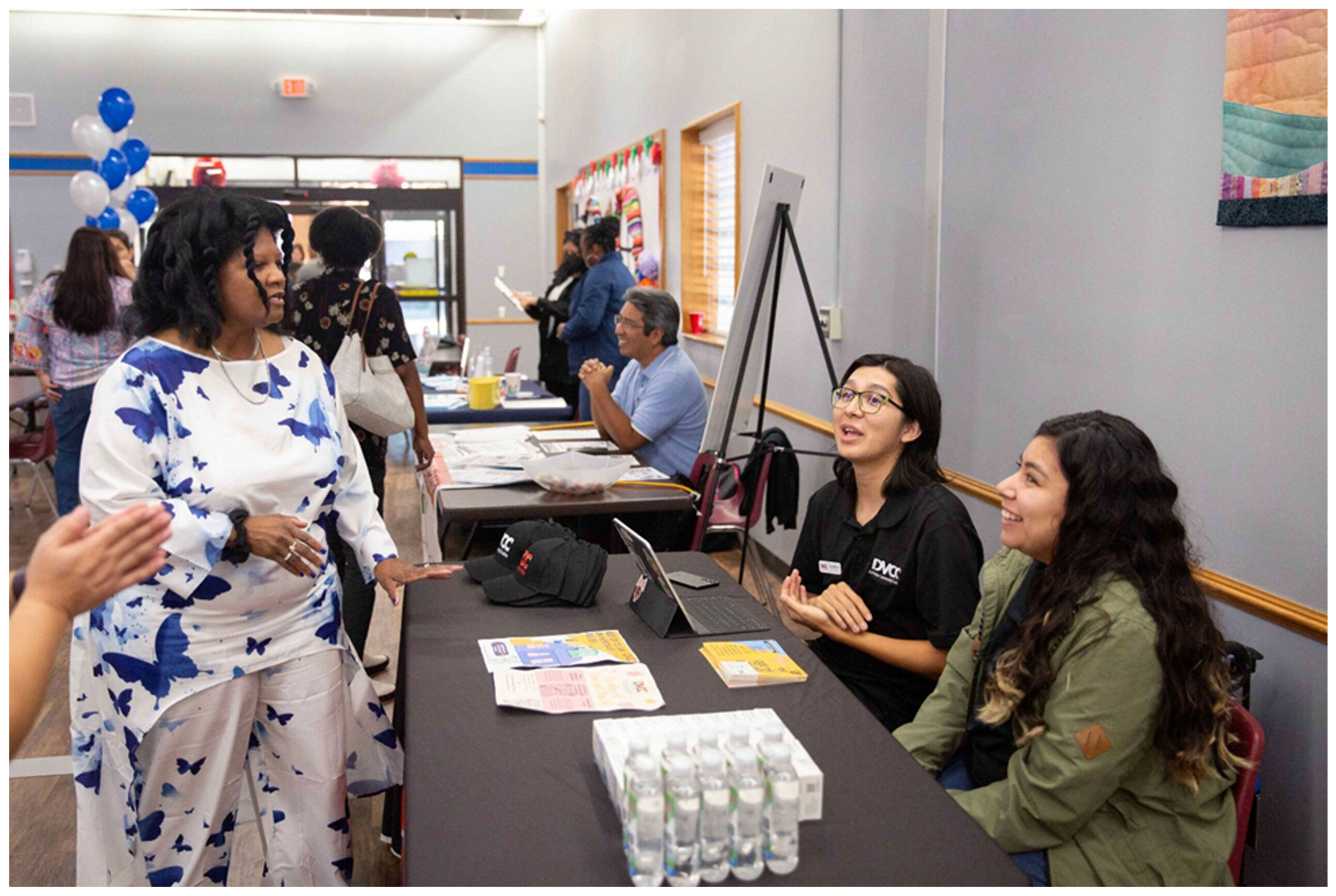 Image shows people seated at tables, providing attendees with information about assistance. In the forefront, two women are seated at a table, speaking with a lady who is standing on the opposite side. There is bottled water on the table as well as informational flyers, hats and a laptop.