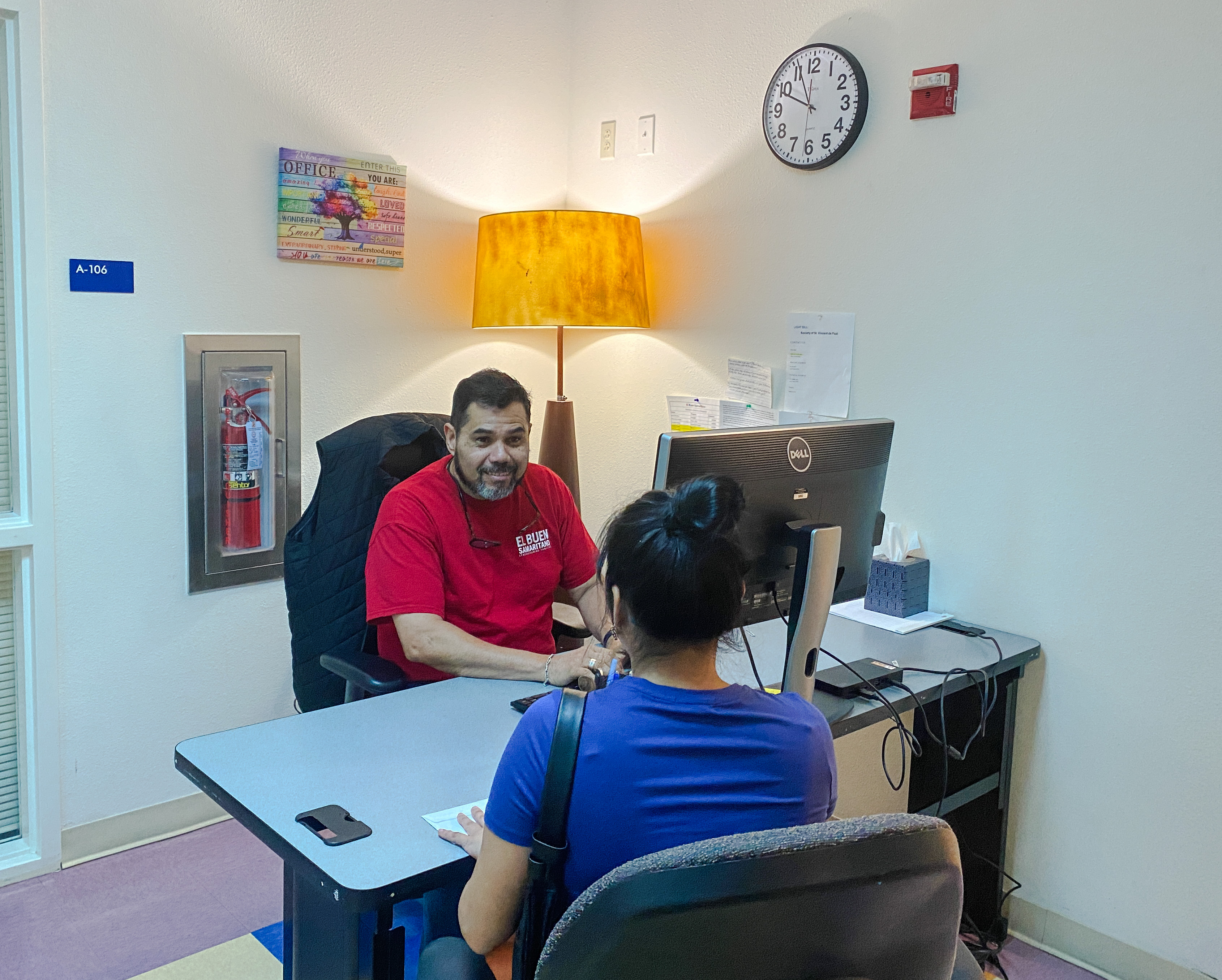 Photo of a male El Buen employee sitting at a desk and assisting a female Austinite, who is sitting on the opposite side of the desk. There is a floor lamp on behind the employee and a clock on the wall..