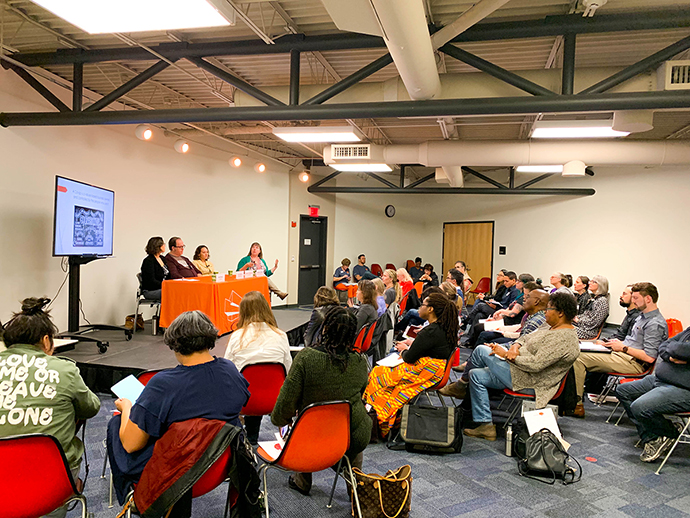 A group of people sitting in red chairs listening to a panel of 4 speakers