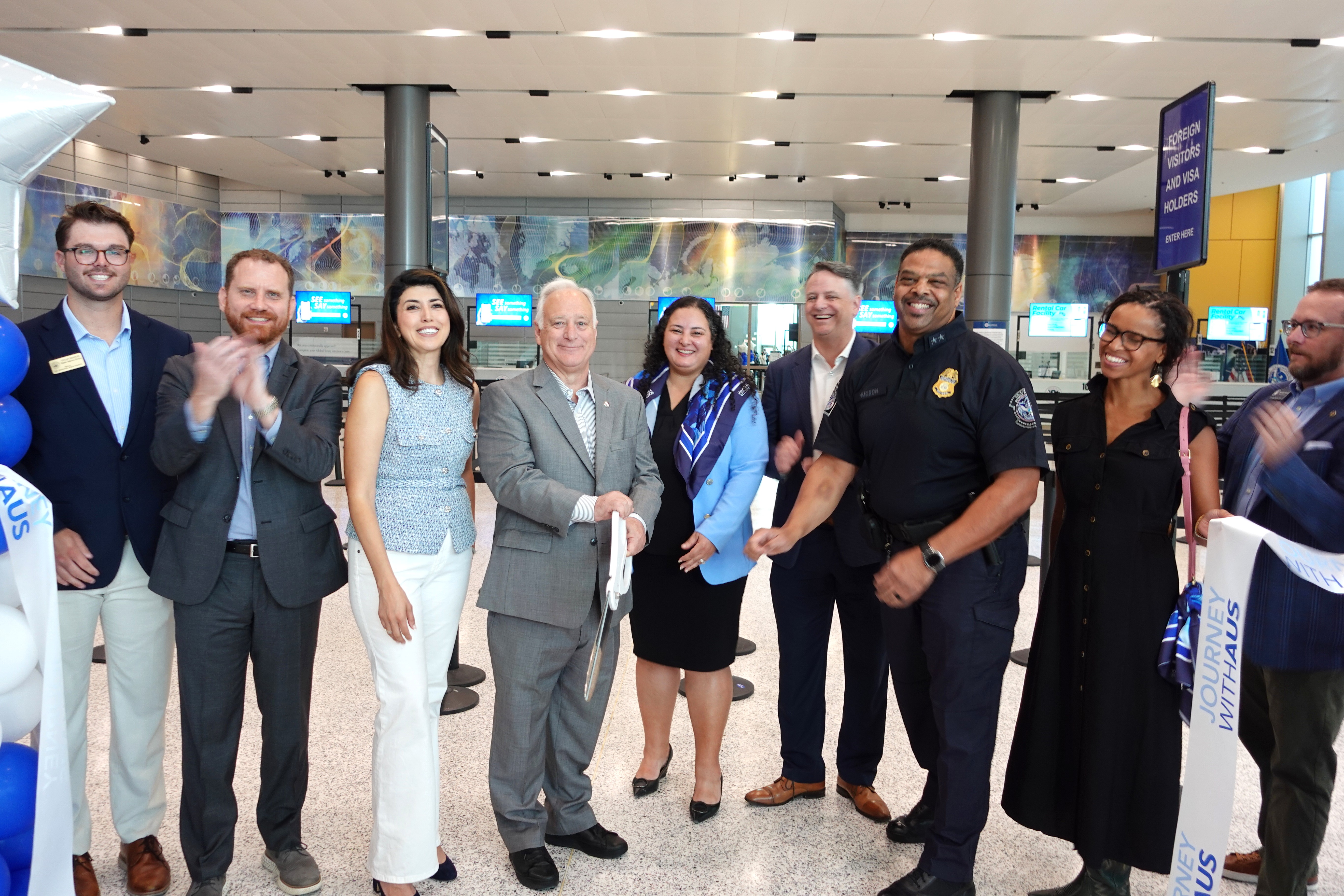 A group of ten people, including airport and government officials, smile and pose during a ribbon-cutting ceremony inside a modern airport terminal. One man in the center holds large ceremonial scissors, while others clap and celebrate. Blue and white balloons and signs reading "Journey With AUS" and "Foreign Visitors and Visa Holders" are visible in the background.