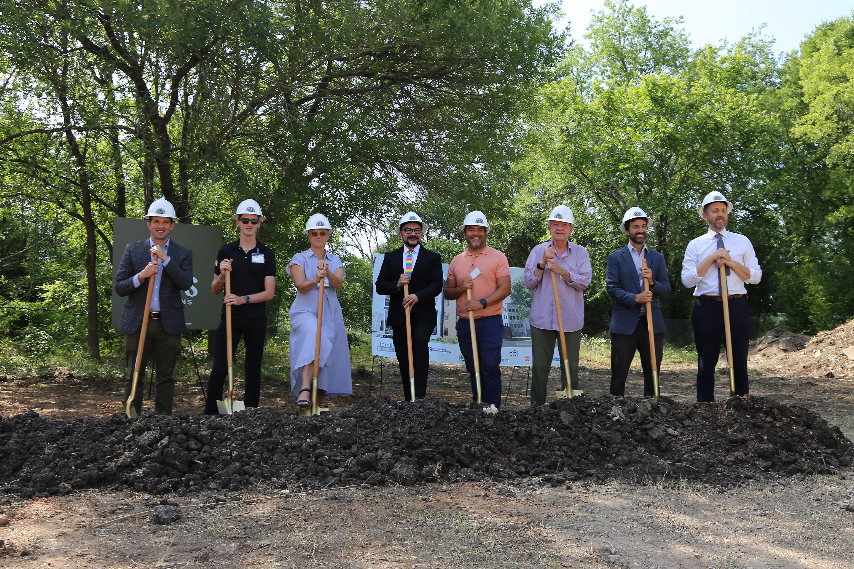 Image shows eight individuals at a groundbreaking. All are standing side-by-side wearing white construction hard hats and holding shovels that are pointed into a mound of dirt.