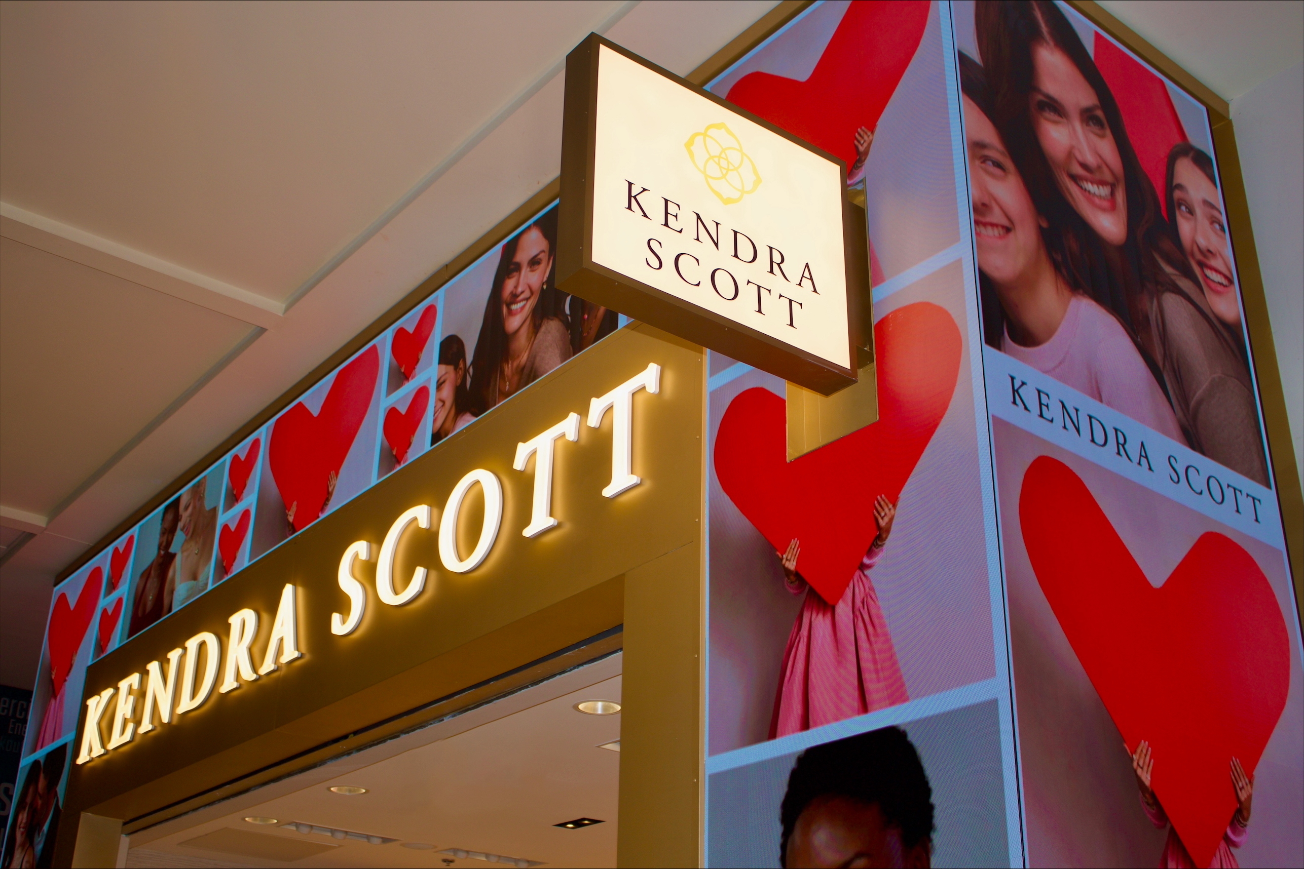 Image of Kendra Scott's airport store. Images of people and red hearts surround an illuminated Kendra Scott logo atop the store's entrance.
