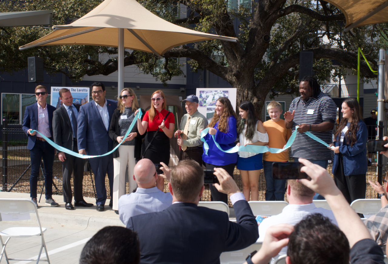 Eleven men, women and children holding a blue ribbon near a children's playground. The man in the center is holding a large pair of red handle scissors.