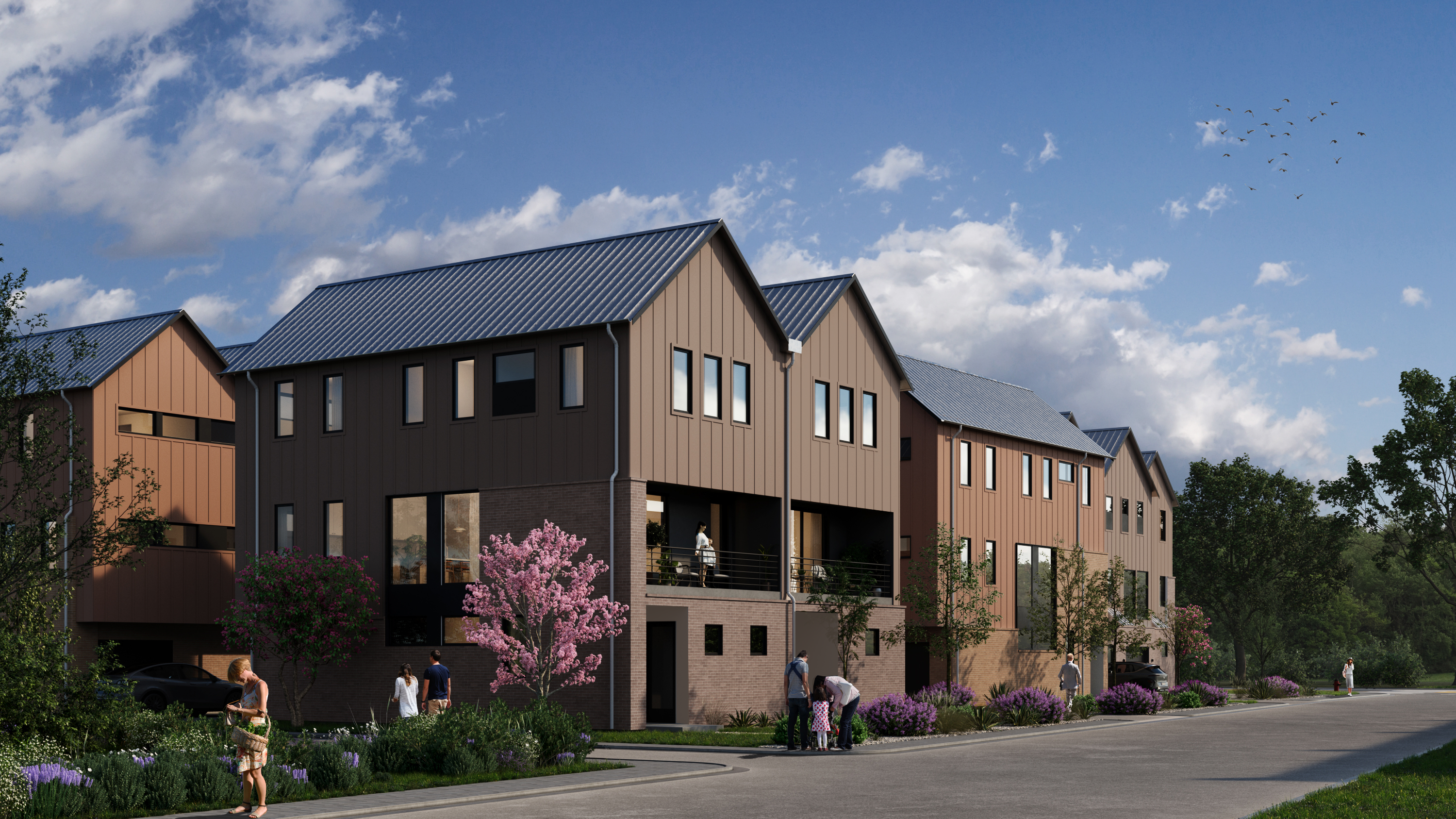 Mira_Springdale-Gonzales Row of modern three-story townhomes with gabled metal roofs and warm-toned siding, surrounded by landscaped greenery, blooming trees, and a clean residential street. Several people are walking or talking outside, contributing to a welcoming neighborhood atmosphere under a partly cloudy blue sky.