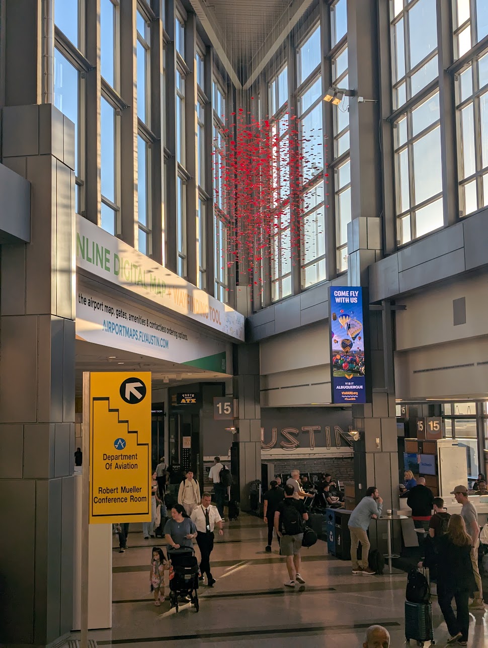 A large-scale red hanging art installation, made up of hundreds of suspended elements, is displayed in the high bay triangle area near Gate 15 inside Austin-Bergstrom International Airport. The installation is positioned against tall glass windows with natural light streaming in, while travelers move through the busy concourse below.