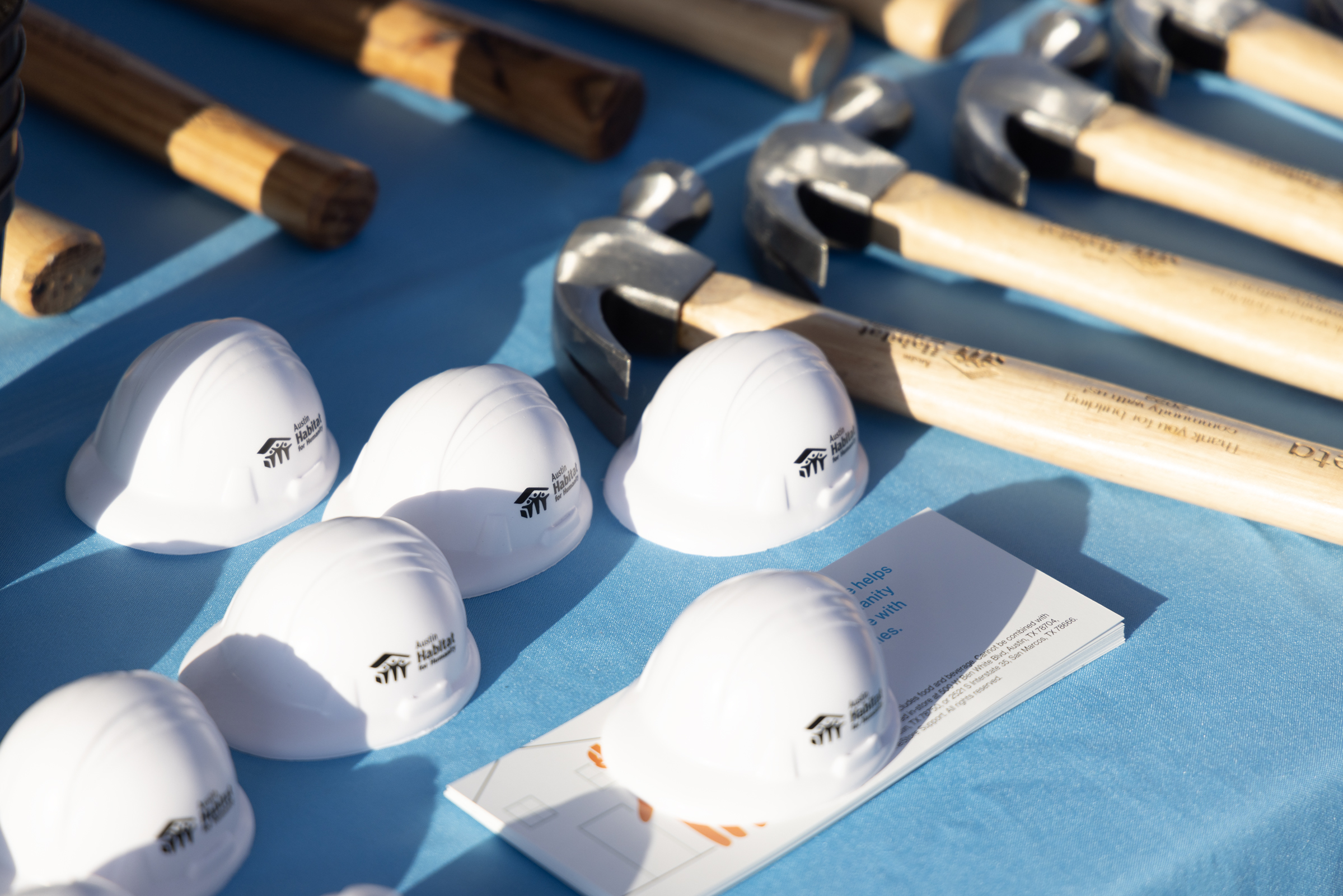Image shows a table covered with a blue tablecloth. On top of the table are several hard hats grouped together along with rows of wood-handled hammers also grouped together.