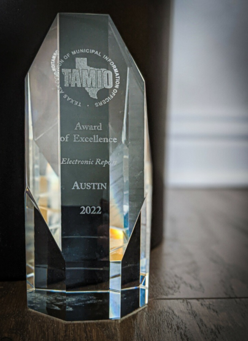 Set against a dark background is the glass cut TAMIO award reading, "Award of Excellence Electronic Report Austin 2022."