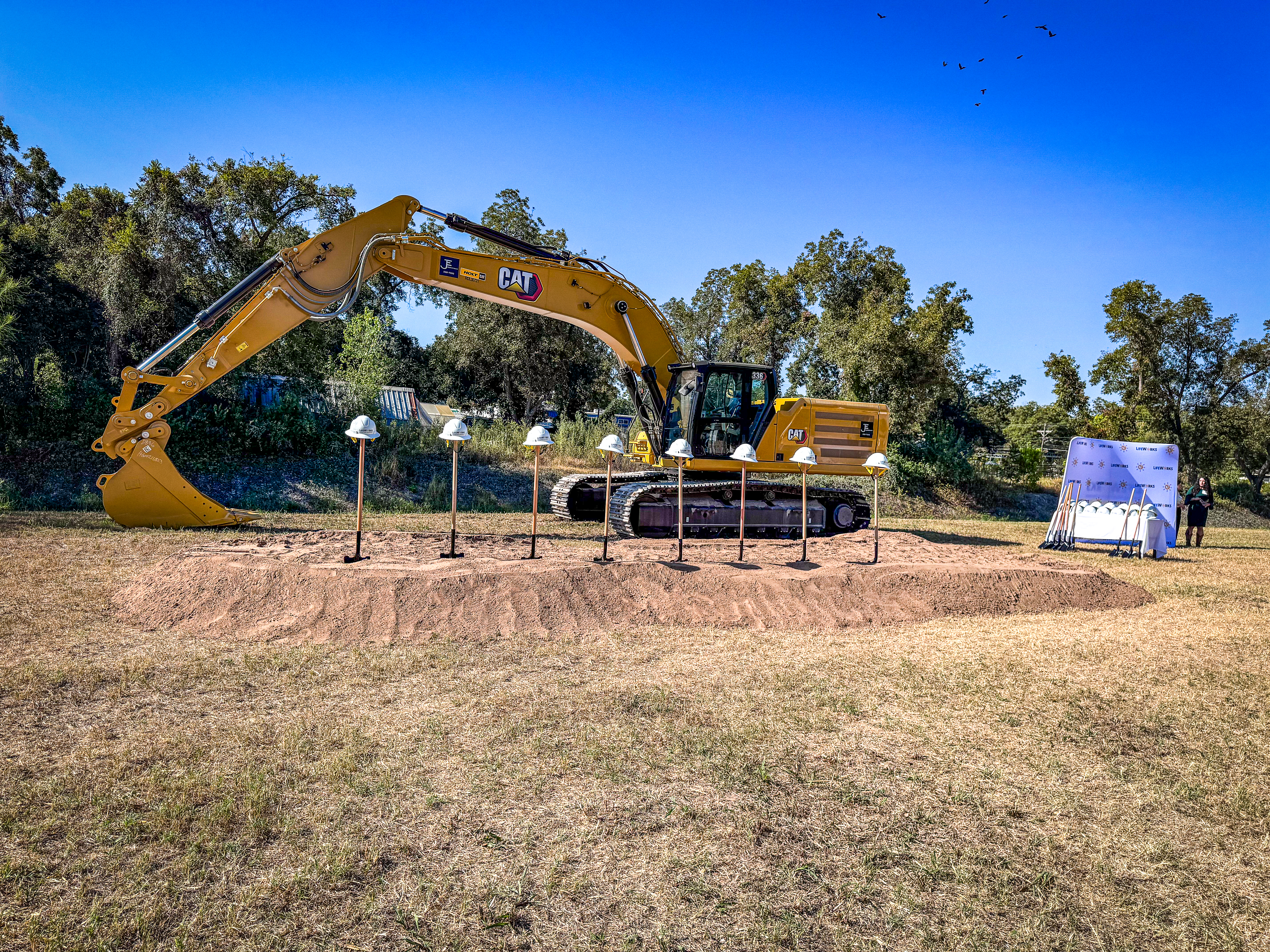 Groundbreaking ceremony setup with a CAT excavator, hard hats on stands, and shovels by a branded backdrop.