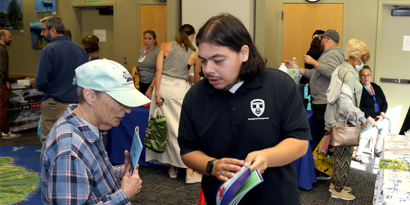 A photo showing a HSEM staff member sharing preparedness information with a community member, while people visit preparedness booths in the background.
