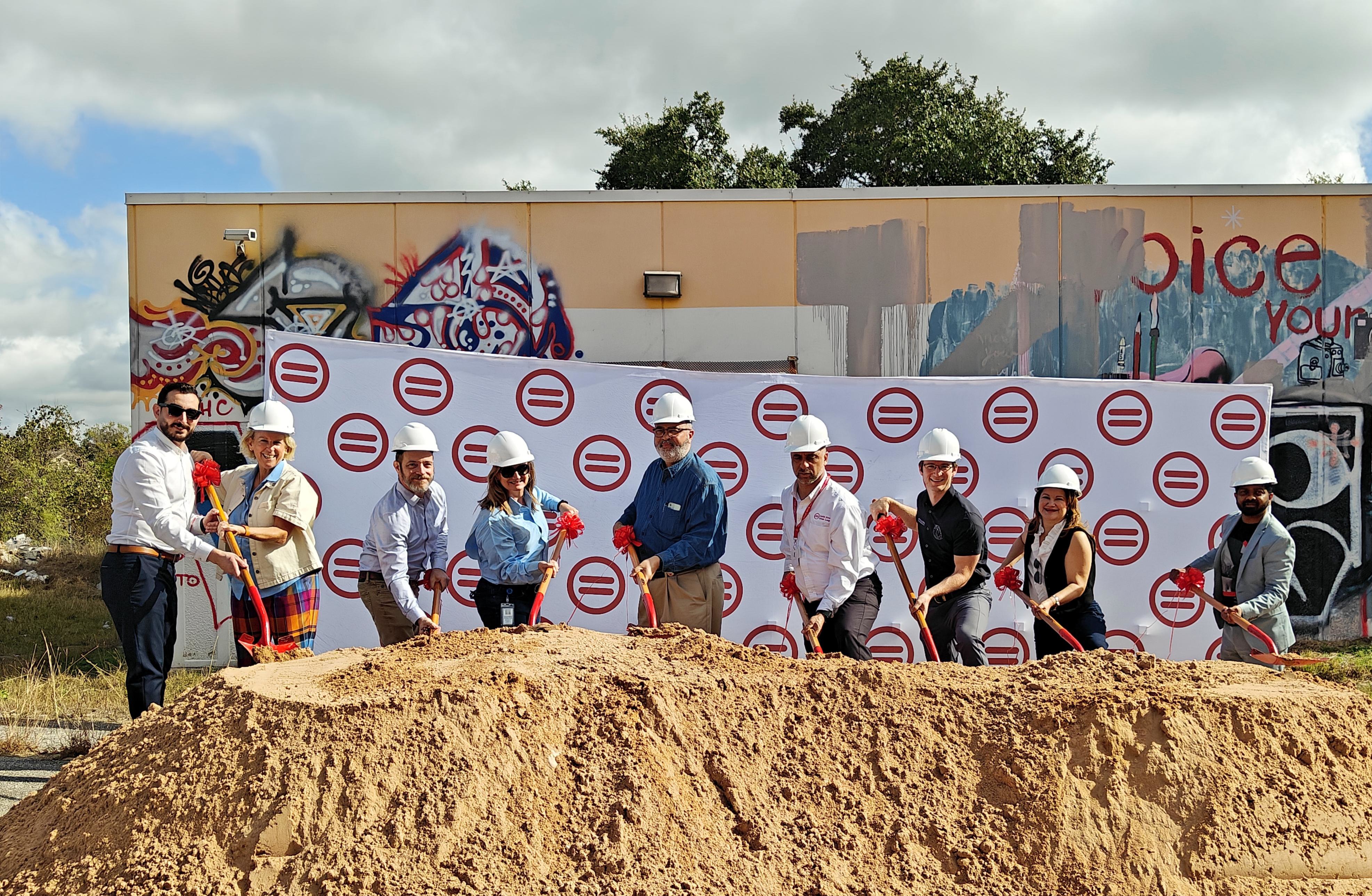 Image shows nine officials, either of which are wearing white hardhats and red holding shovels, standing behind a large mound of dirt. A graffitied trailer is behind them. 