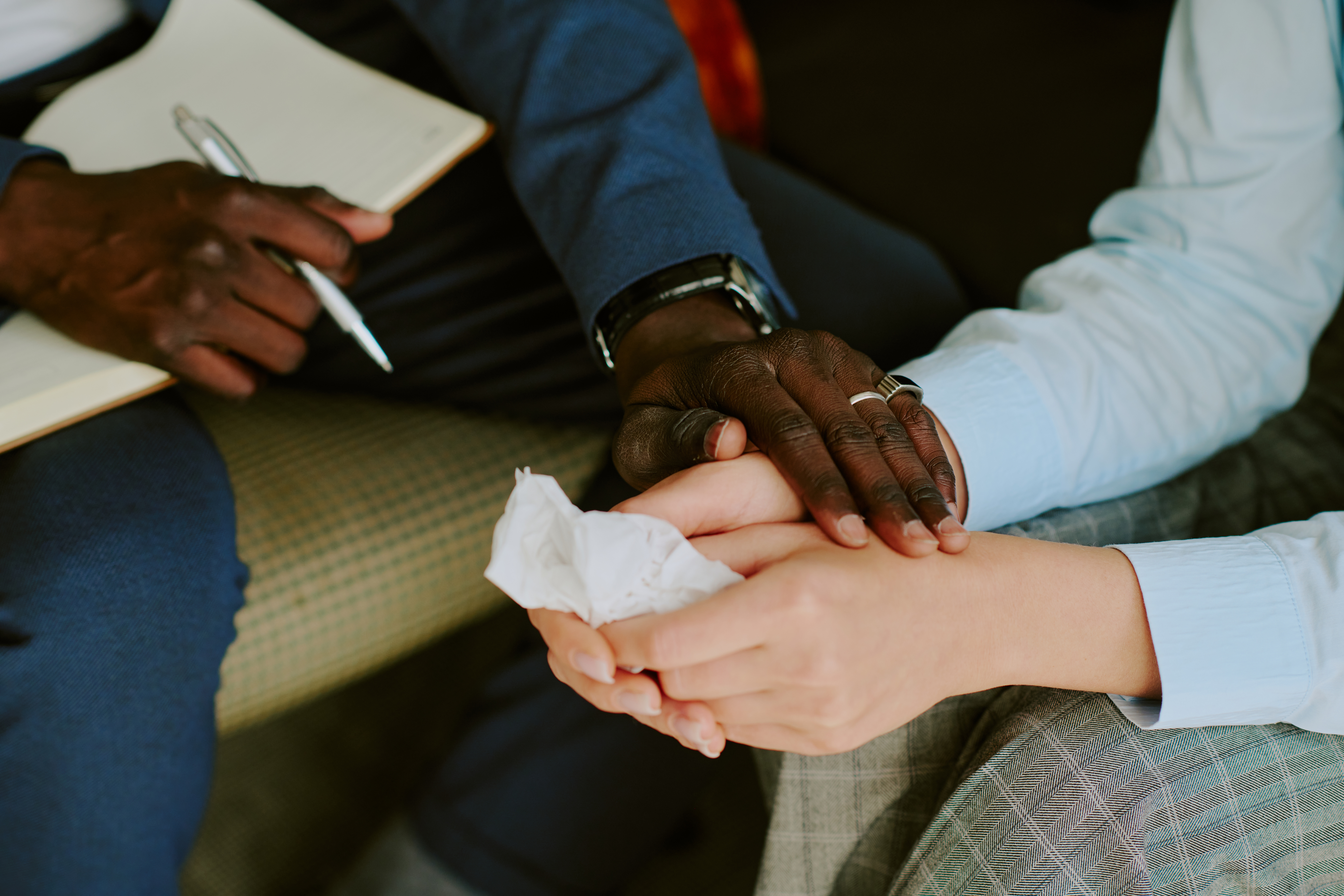 tight photo of a black man's and white woman's hands. The man holds a notepad and pen in one hand and holds the woman's hands with the other. She holds a facial tissue.