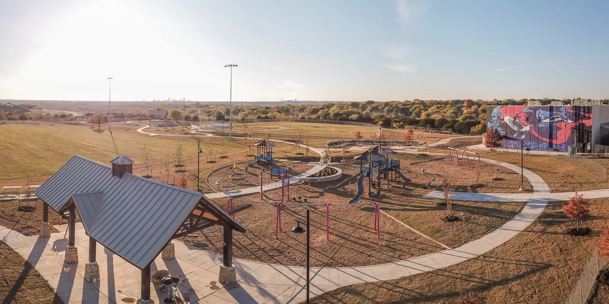 Image shows a park in Colony Park with a covered area, swings and slides in the foreground and a community center in the background with a blue, white and pink mural painted on the back of it.