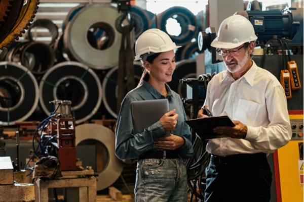 A woman and man wearing hard hats stand in a warehouse discussing material written on a clipboard