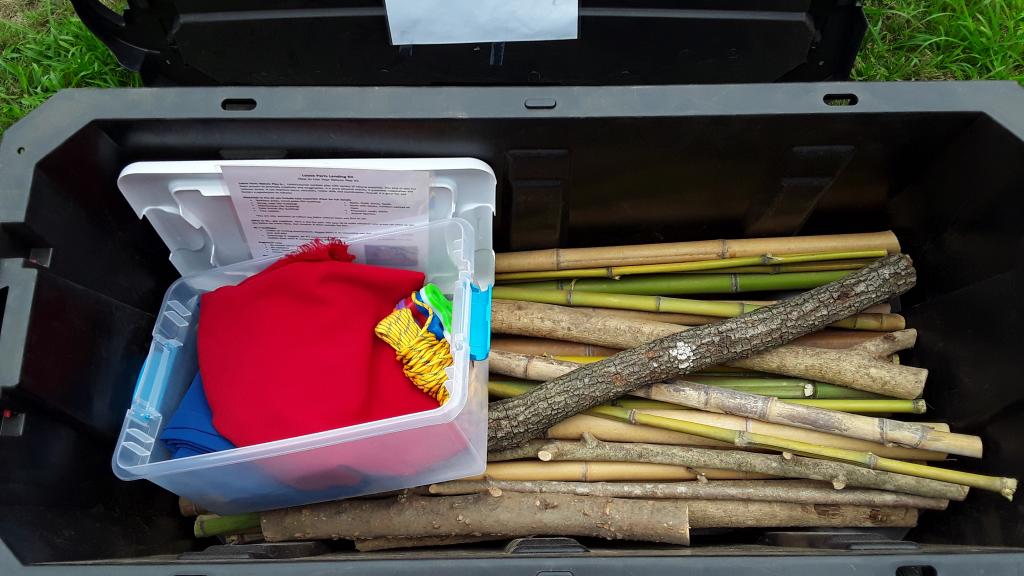 A large, black storage container containing bamboo sticks and tree branches. Resting on top of the bamboo and tree branches is a smaller, translucent box containing fabric pieces and rope.