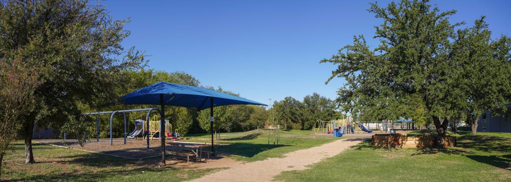 A panaramic view of the current playground with shade structure over a picnic table and playscapes.