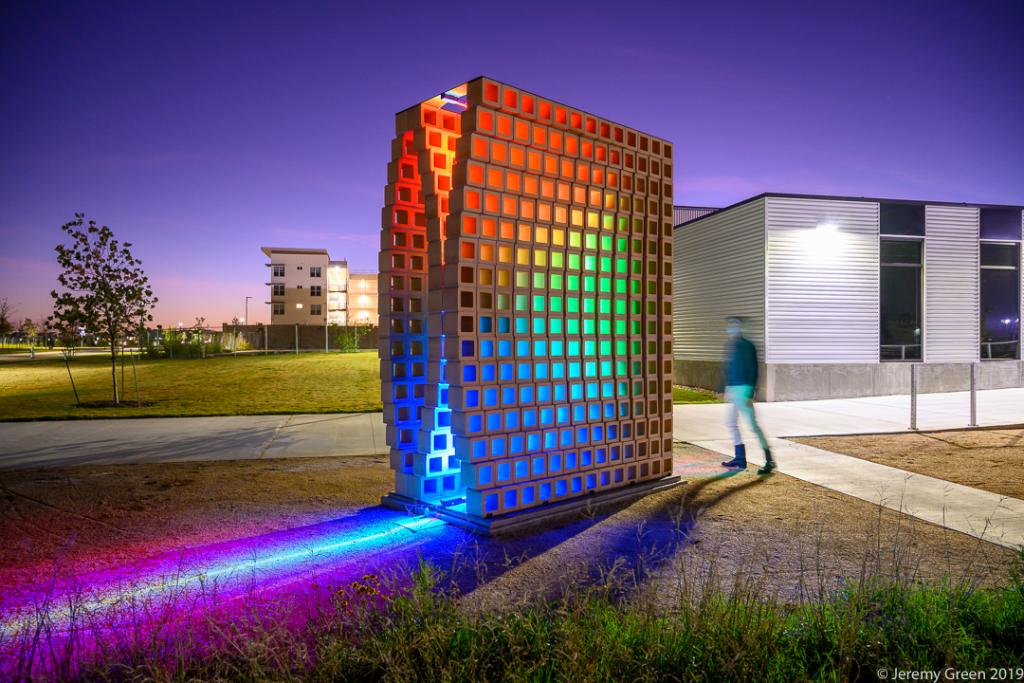 Illuminated public art installation made of a grid-like structure glowing in rainbow colors at dusk, with a person walking past nearby buildings.