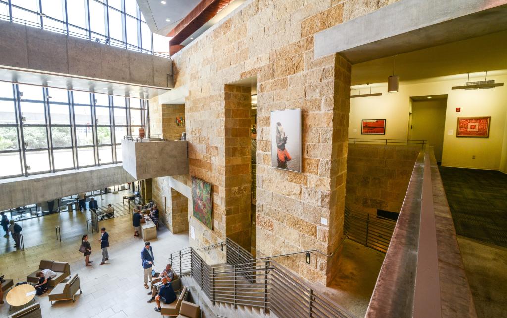 Interior view of a large, light-filled gallery space with tall stone walls, concrete walkways, and floor-to-ceiling windows. Framed artworks hang on the walls along multiple levels. Visitors gather and converse on the ground floor seating area and near a reception desk, while staircases and railings connect the open, multi-story interior.