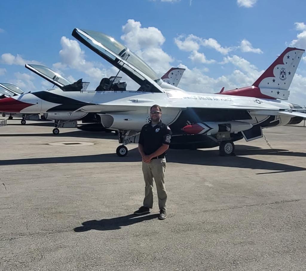 A man wearing sunglasses and a black polo with khaki pants stands in front of a row of United States Air Force Thunderbirds fighter jets on a sunny day. The aircraft are parked on a tarmac, and the sky is bright blue with scattered clouds.