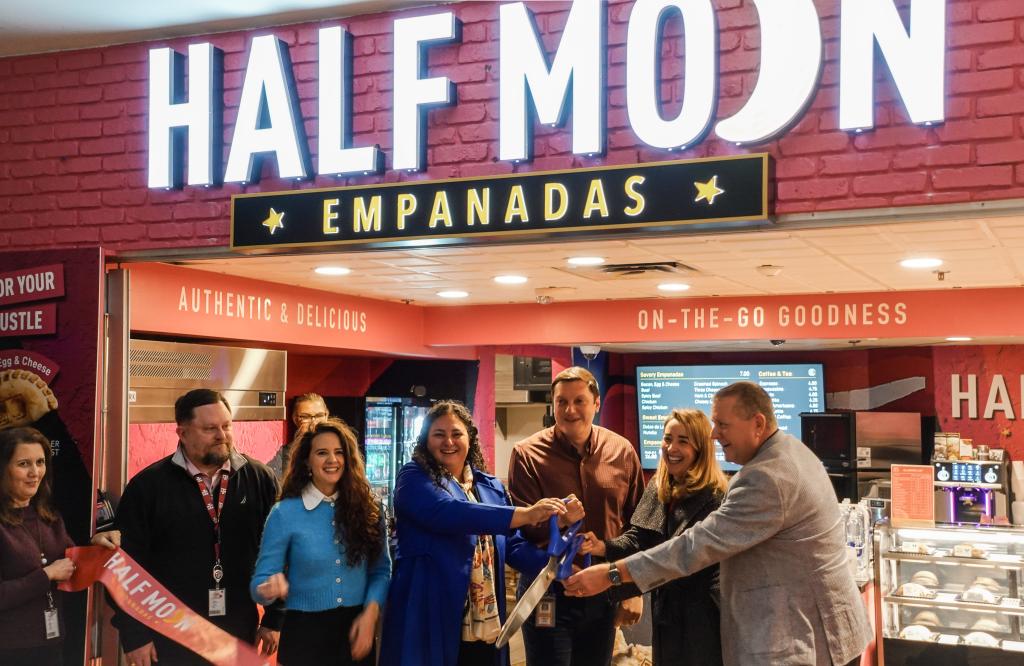 A group of people gathers in front of the Half Moon Empanadas storefront for a ribbon-cutting ceremony. The sign above the shop is brightly lit, with "EMPANADAS" highlighted below. The interior features a red and white color scheme with signage reading "Authentic & Delicious" and "On-The-Go Goodness." The individuals, wearing a mix of business and casual attire, are smiling as they cut the ceremonial ribbon with large scissors, marking the grand opening of the new food outlet.