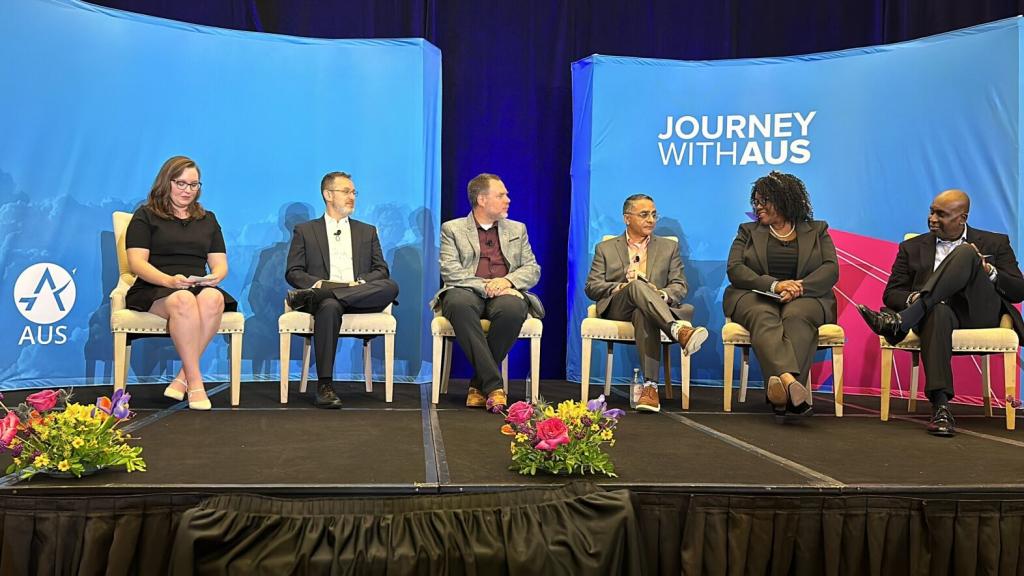 Six panel members sitting in chairs on a stage with a blue background that have the Austin airport logo and Journey With AUS brand.