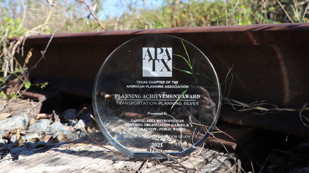 Photo of a clear, glass award that reads Planning Achievement Award positioned in front of a brown steel rail.