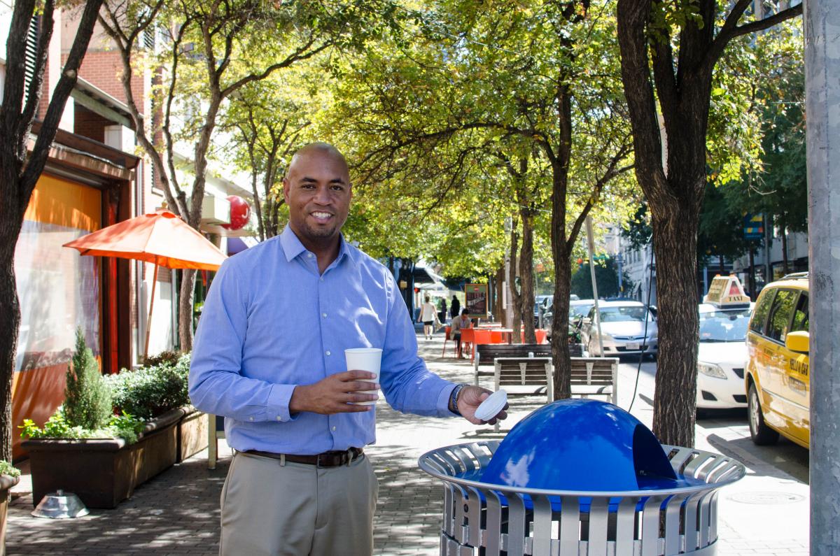A photo of August recycling the top of his coffee cup in a blue recycling bin,