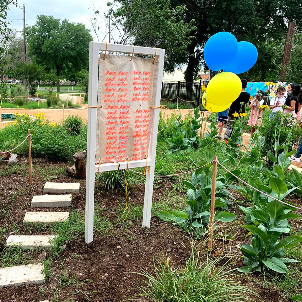 Photo of a carden. Grass and several flowers with long stalks grow surrounding a canvas sign with the words "love each other" repeating in a script font.