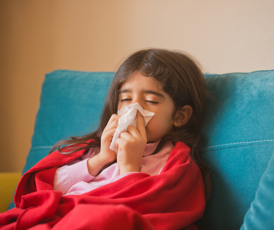 A young girl with a blanket on a couch holding a tissue to her nose.