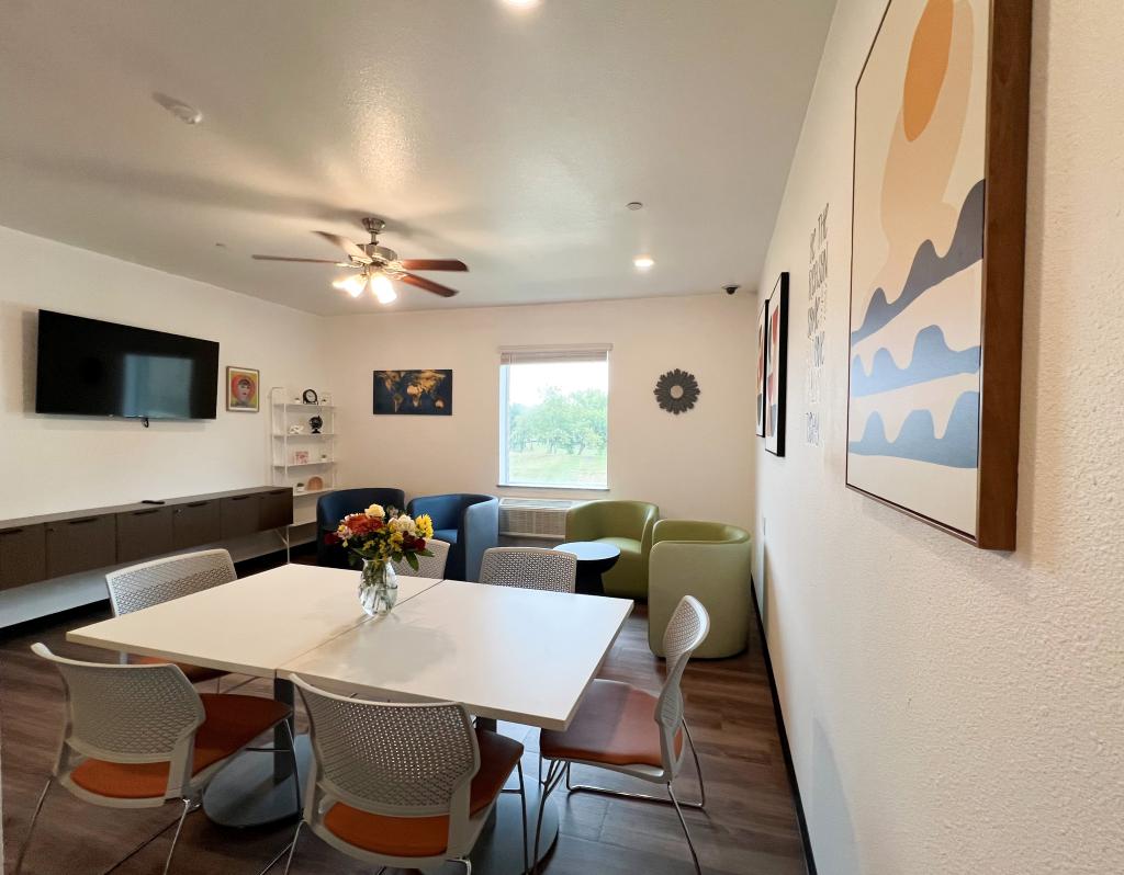 Image shows a white square dining table for six, a large television mounted on the wall with a floating entertainment center, 2 blue and 2 green curved chairs and wooden side tables.