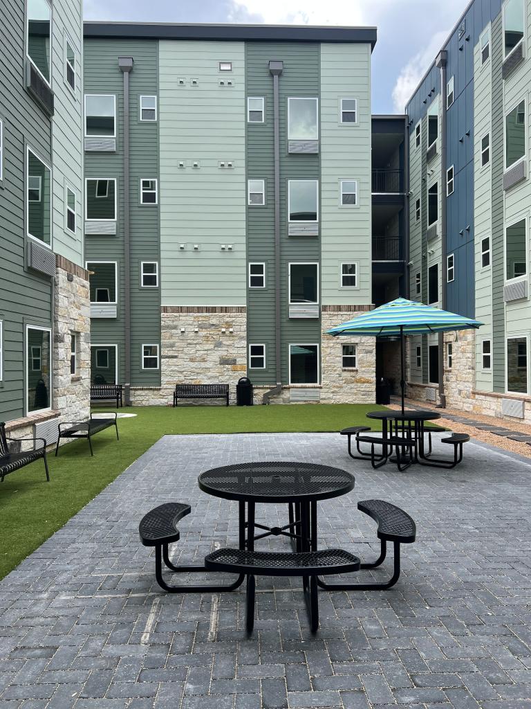 Image shows inner courtyard of the Cady Lofts apartments. The courtyard is surrounded by tall multi-storied buildings that have a stone and blue and green siding exterior. The rectangular courtyard has several benches on the outer edges with two tables with benches on a brick-paved surface. 