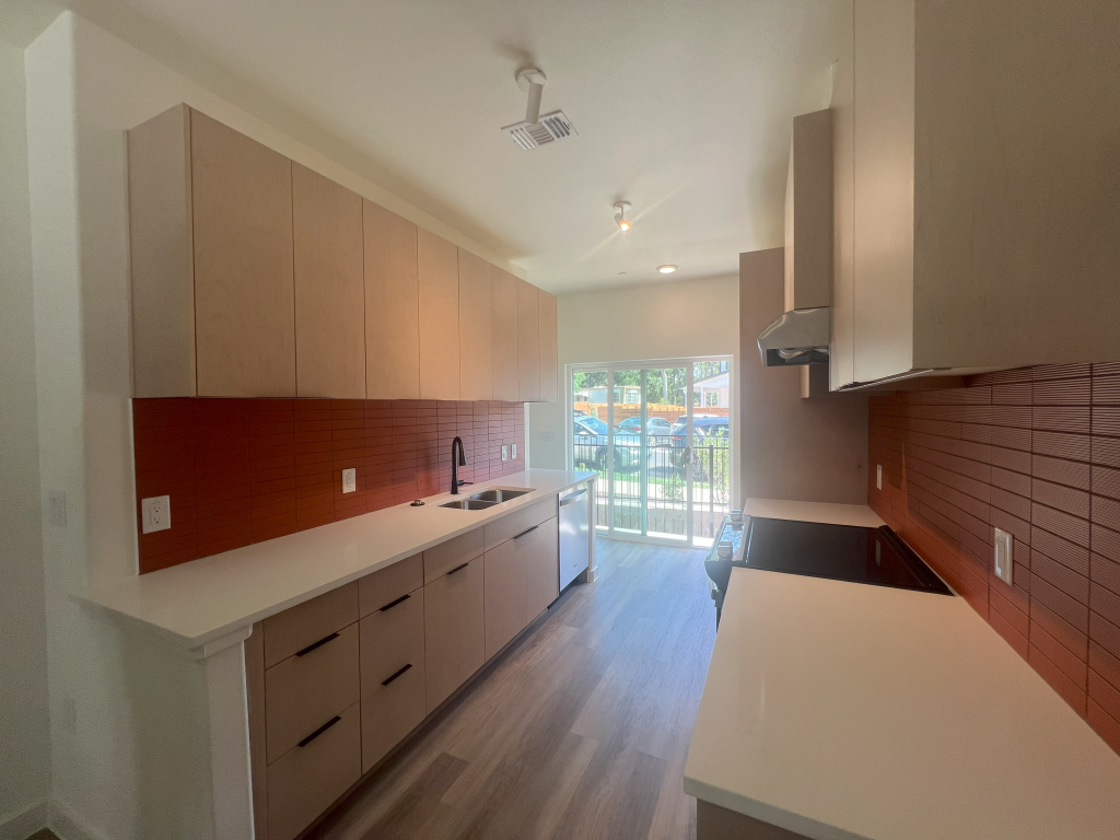 Image shows view of kitchen in four-bedroom townhome. Cabinets are in a light wood finish and there is a red horizontal tile backsplash and white quartz countertops.